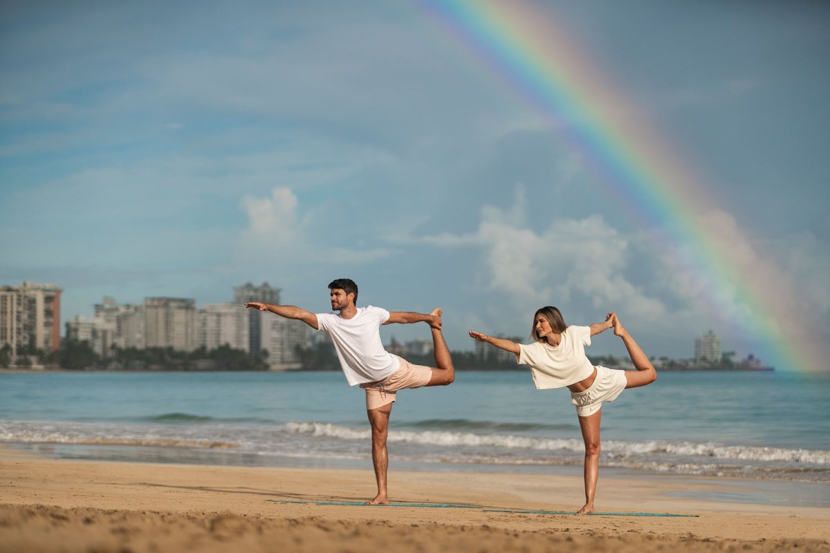 A couple practices yoga on a beach with a rainbow overhead at Fairmont El San Juan hotel.
